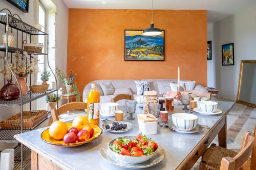 Salle à manger lumineuse du Jardin Secret Grignan avec mur d'accent orange, petit-déjeuner garni de fruits frais et viennoiseries