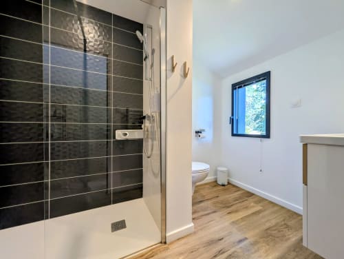 Salle de bain moderne à La Clairière aux Chênes avec douche en carrelage noir, toilettes blanches, parquet et fenêtre bleue sur jardin