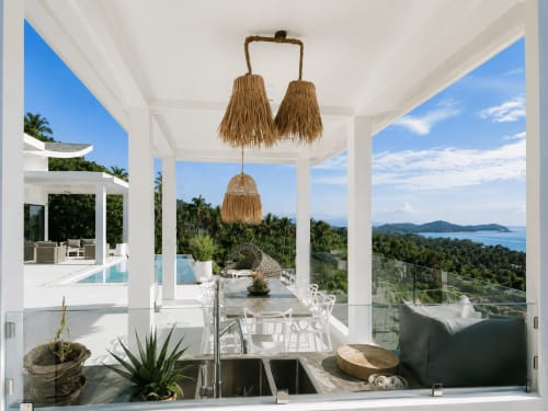 Terrasse de villa moderne avec vue sur la mer à Koh Samui, table de salle à manger, luminaires suspendus tissés et paysage tropical.