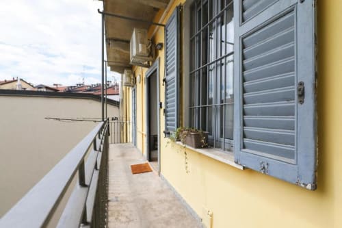 Stretto balcone di appartamento milanese con muri gialli, finestre con persiane blu e vista sui tetti della città