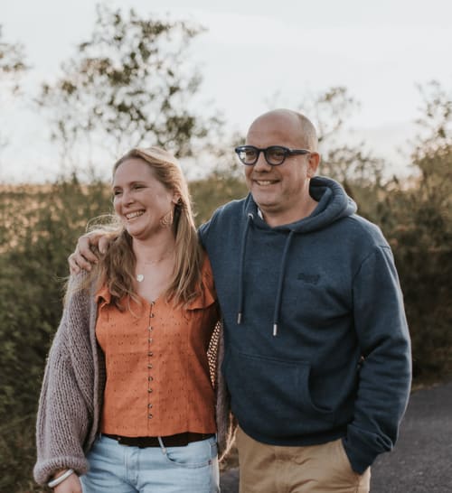 Couple souriant en extérieur aux Gîtes du Bulz, une chambre d'hôtes à Pleyber-Christ, France, avec un paysage forestier derrière eux.