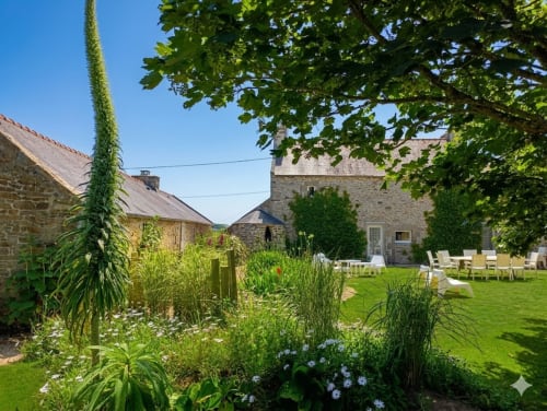 Jardin du gîte Ker Gana avec bâtisses en pierre, pelouse verdoyante, fleurs épanouies et arbre ombragé à Plourin, France.