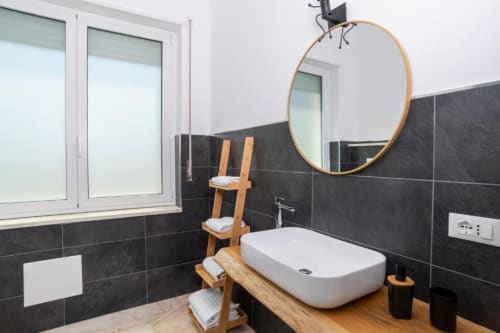Bagno moderno presso il Domo Cadò B&B a Berchidda con lavabo bianco a bacinella, specchio rotondo, piastrelle nere, scala in legno e