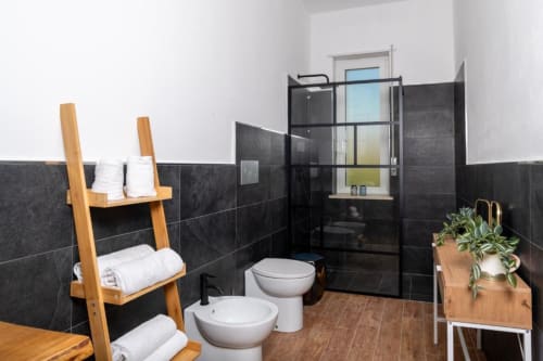 Bagno moderno presso Domo Cadò BnB a Berchidda con piastrelle nere, sanitari bianchi, porta asciugamani in legno e vista sul giardino