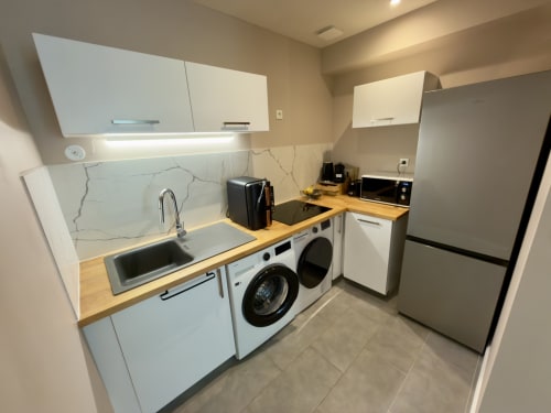 Modern kitchenette in Castres apartment with white cabinets, wooden countertop, sink, washing machine, and stainless steel