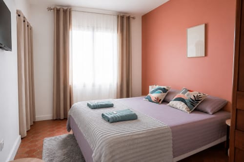 Chambre moderne avec mur d'accent corail, lit queen avec coussins décorés, rideaux beige et vue sur le jardin au Mas De La Clé