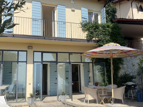 Patio der Villa Casa Aurora in Salò mit gestreiftem Sonnenschirm, Essstühlen und modernen Glastüren mit Blick auf den Gardasee