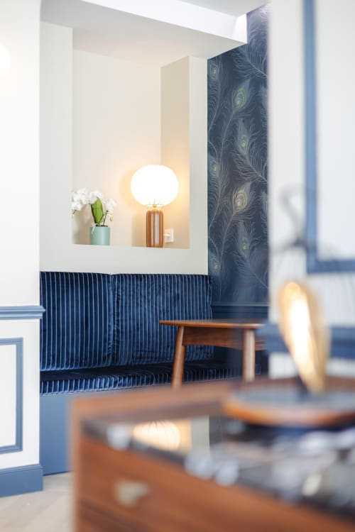 Le salon L'Ode à Pierre BnB avec sièges bleu marine côtelés, papier peint paon et lampe de table allumée, Dax