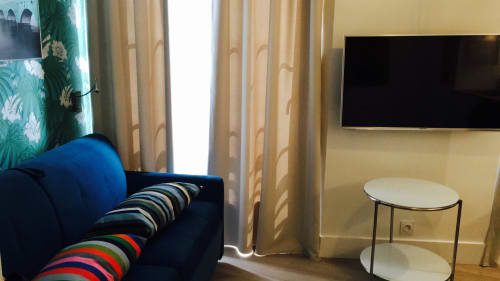 Modern studio at Cocoon Gallery Paris with blue sofa, striped cushions, flat-screen TV, and round side table by curtained