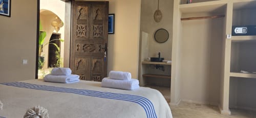 Chambre Mogador du Riad Arkaya à Essaouira : literie blanche, serviettes roulées, porte en bois sculptée et vue sur le jardin