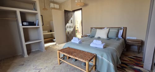 Chambre Enakhla du Riad Arkaya à Essaouira : lit queen-size, banc tissé, étagères et porte ouvrant sur le jardin