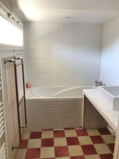 Salle de bain moderne du gîte Les Petits Toits avec carrelage blanc, sol à damier rouge et crème, baignoire, lavabo et fenêtre.