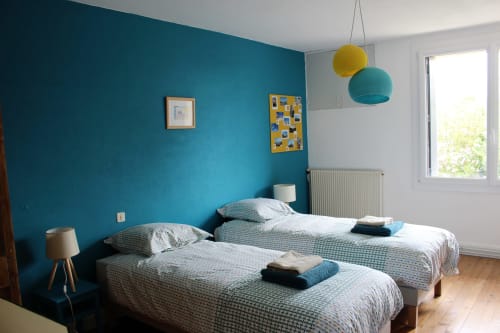 Lits jumeaux avec accents bleu-vert à la gîte Les Petits Toits, Vérac, avec mur de couleur bleu et luminaires suspendus