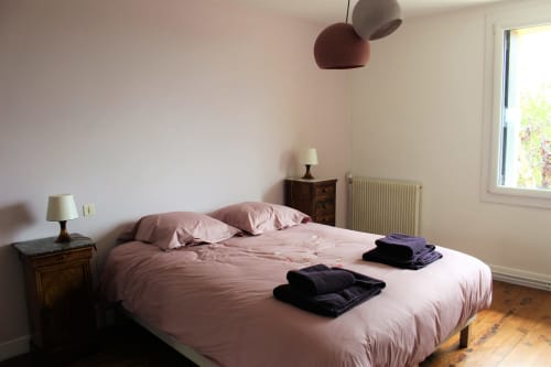 Chambre douillette avec literie rose, table de nuit en bois foncé, lampe suspendue et vue sur le jardin au gîte Les Petits Toits à Vérac