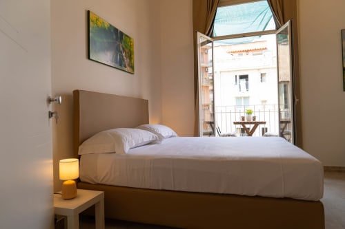 Bright bedroom with queen bed, white linens, tan headboard, open balcony doors overlooking Catania cityscape, modern