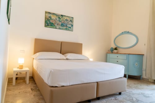 Bright bedroom in Sicily In Home apartment with beige bed, white linens, turquoise dresser, and artwork