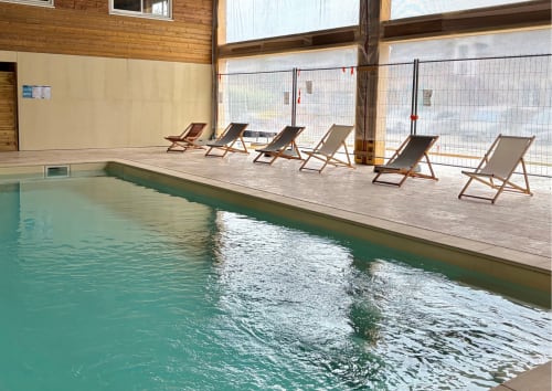 Piscine intérieure du Domaine Les Racines avec eau turquoise, terrasse en bois, chaises longues et baies vitrées panoramiques