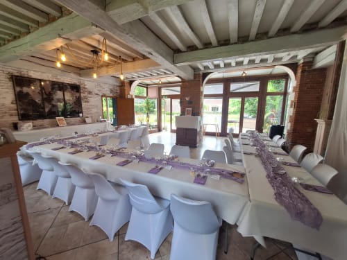Salle de banquet élégante à La Cour d'Hortense avec table en U, housses blanches sur les chaises, accents violets et poutres apparentes.
