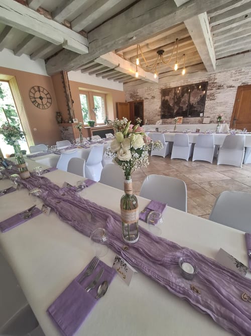 Élégante salle de réception à La Cour d'Hortense avec chemins de table violets, chaises blanches et poutres apparentes