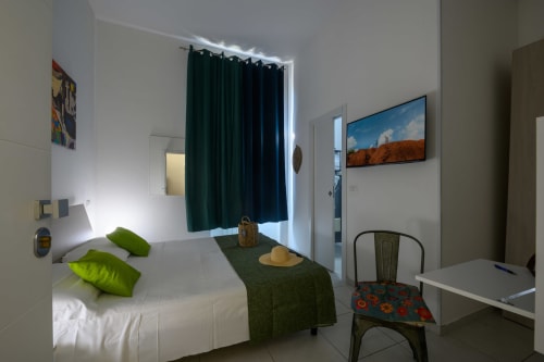 Modern hotel room at Dimore Salentine in Lecce with bed, green pillows, dark curtains, TV, and desk