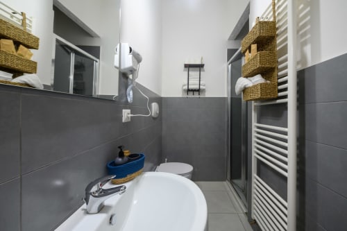 Modern bathroom at Dimore Salentine in Lecce with gray tile walls, white sink, shower, and woven storage baskets