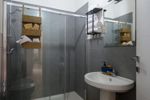 Modern bathroom at Dimore Salentine in Lecce with walk-in shower, white sink, woven storage baskets, and gray tile walls