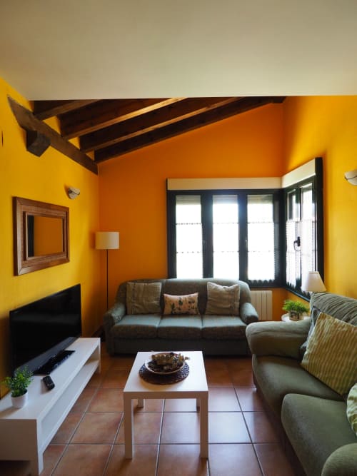 Luminosa sala de estar con paredes en naranja y amarillo vibrante, techo con vigas oscuras, sofá gris, televisor y grandes ventanales con vista al jardín