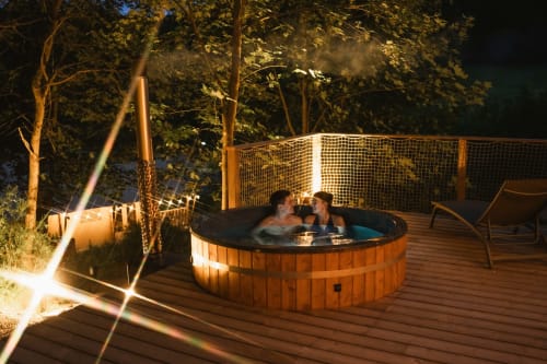 Couple se relaxant dans un bain à remous en bois sur la terrasse des Cabanes du Nideck, Oberhaslach, entouré par l'éclairage forestier nocturne