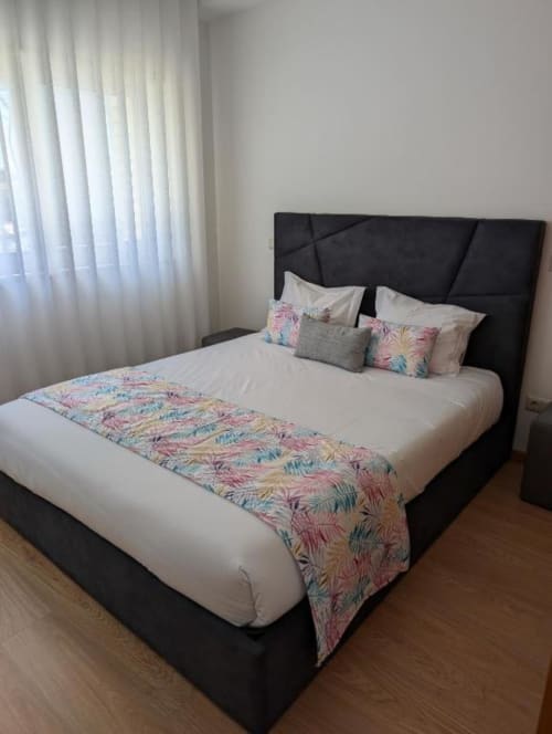 Quarto moderno em Villas Eden com cama estofada preta, roupa de cama branca, manta colorida estampada e cortinas translúcidas
