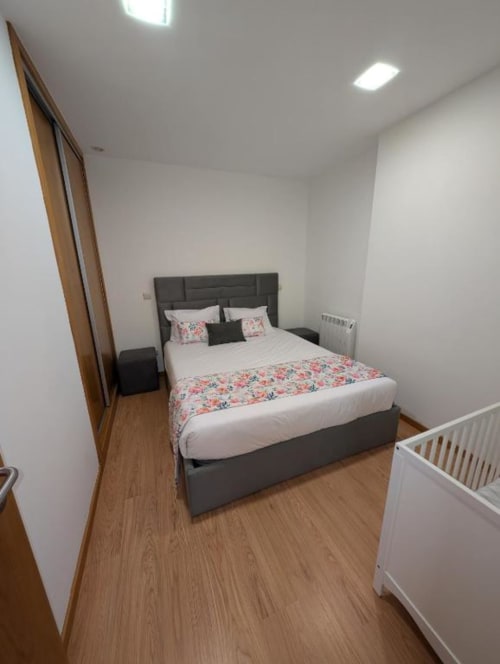 Quarto moderno com cama cinza, roupa de cama floral, berço branco, piso de madeira e iluminação embutida no Villas Eden