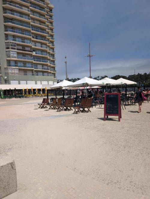 Espaço de bar praia com guarda-sóis brancos, cadeiras castanhas e painel de menu vermelho em Villas Eden em Fão, Portugal, com apartamento moderno