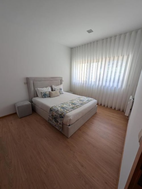 Quarto luminoso no apartamento Villas Eden com cama bege, cortinas brancas translúcidas, piso de madeira e vista para o jardim