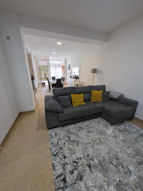 Sala de estar moderna no apartamento Villas Eden com sofá cinza, almofadas amarelas, tapete estampado e cozinha à vista
