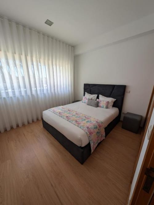 Quarto moderno na Villas Eden com cama preta, roupa de cama branca, caminho floral, cortinas brancas, piso de madeira e vista para o jardim