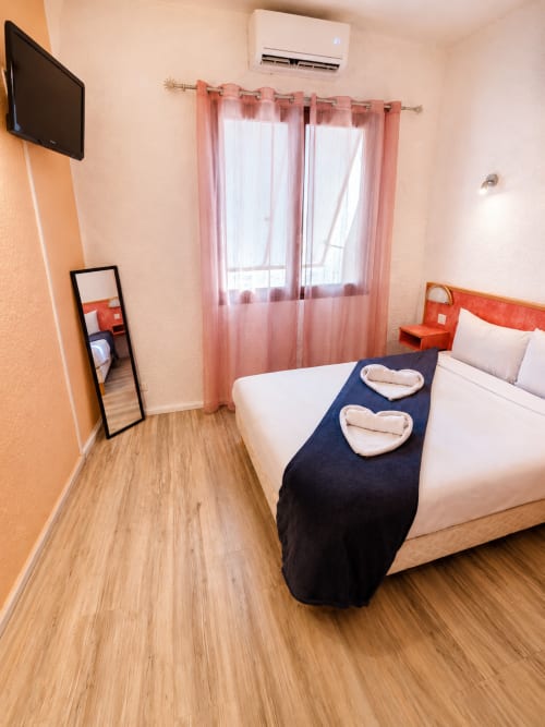 Chambre double à l'Hôtel Les Alizés au Pouzin avec lit blanc, rideaux roses, télévision et parquet