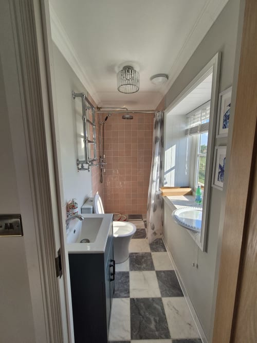 Narrow cottage bathroom at Rose Vill Homes with pink tile shower, white sink, toilet, checkerboard floor, and bright window