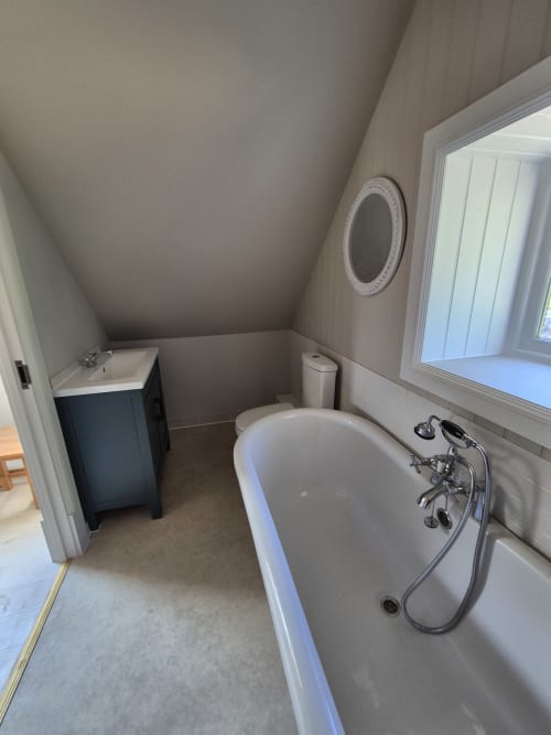 Attic bathroom in Rose Vill Homes cottage with freestanding bathtub, pedestal sink, toilet, and circular window overlooking