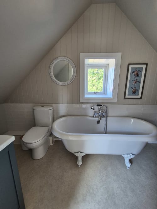 Attic bathroom at Rose Vill Homes cottage featuring a clawfoot bathtub, toilet, round mirror, and garden view window