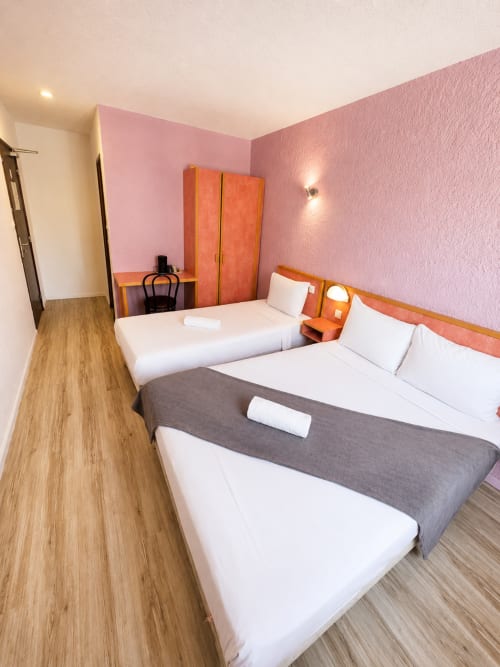 Chambre triple de l'Hôtel Les Alizés avec deux lits, murs roses, mobilier en bois orange et parquet à Le Pouzin
