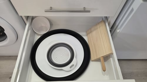 Cajón de cocina abierto con sartén negra, colador blanco, tabla de cortar de madera y recipiente de plástico en MONICA SOCARICI