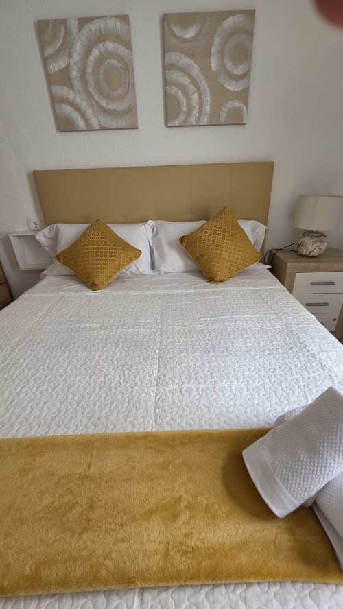Dormitorio de cabaña frente al mar con almohadas mostaza, ropa de cama blanca, cabecero tapizado y arte de pared moderno en Monica Socarici