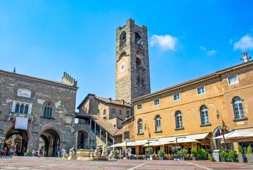 Piazza storica di Treviglio con torre dell'orologio in pietra, edifici medievali e tavoli all'aperto sotto ombrelloni