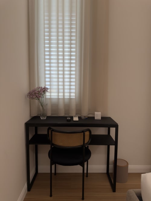 Spazio di lavoro minimalista a Maison Riviera: scrivania nera, sedia in rattan, fiori secchi e luce naturale soffusa dalle persiane