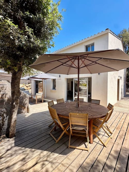 Terrasse moderne avec terrasse en bois, table ronde sous parasol taupe, chaises longues, maison blanche, ciel dégagé