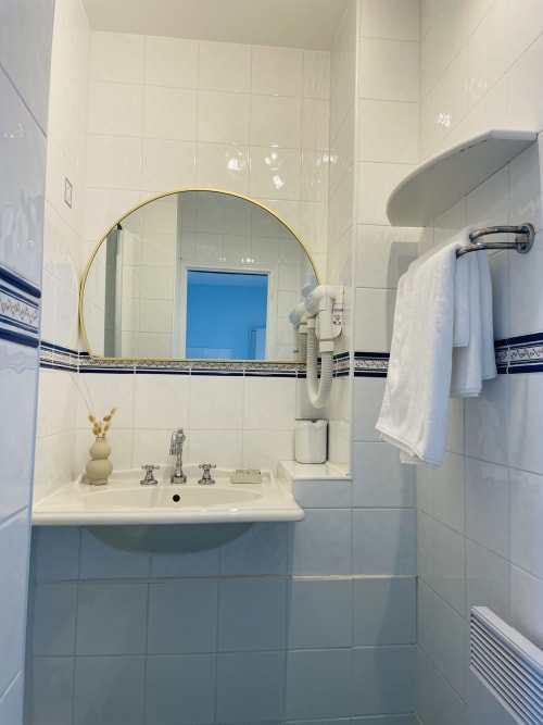 Salle de bain moderne avec miroir rond encadré or, carrelage blanc, lavabo sur piédestal et porte-serviettes à la Maison Nolette B&B