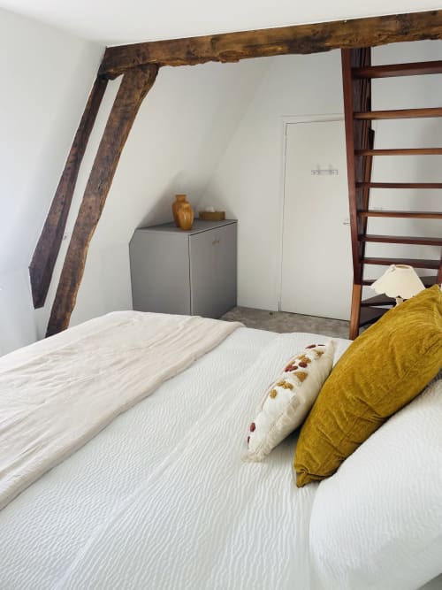 Chambre cosy à la Maison Nolette avec poutres apparentes, literie blanche, coussins dorés et meuble de rangement gris