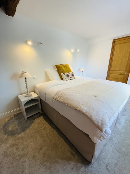 Chambre lumineuse à la Maison Nolette avec literie crème, coussin moutarde, table de nuit blanche et porte en bois