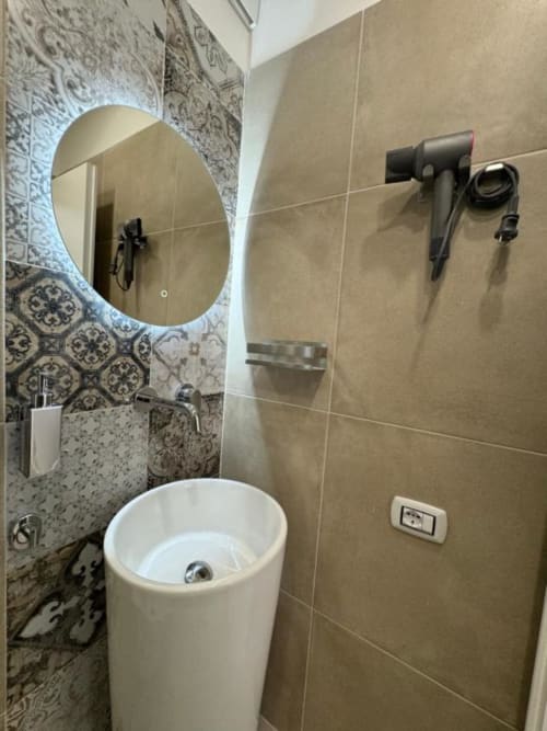 Bagno moderno della Maison ì con lavabo bianco a colonna, specchio rotondo illuminato, parete con piastrelle decorate e asciugacapelli nero