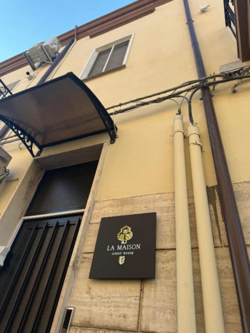 Facciata esterna di La Maison Guest House a Reggio di Calabria con fronte color crema, porta d'ingresso con baldacchino nero e lampade decorative
