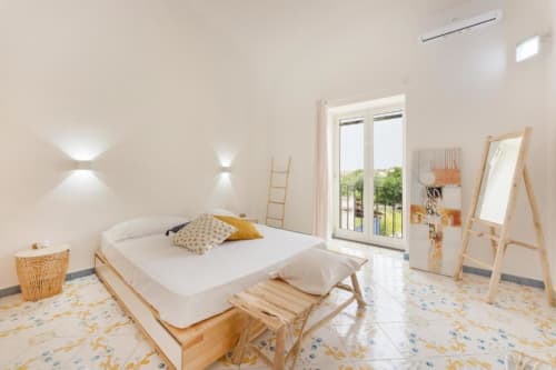 Suite camera moderna e luminosa all'IL POETA di Procida con letto in legno, arredamento minimalista e porte-finestre sulla terrazza con vista giardini
